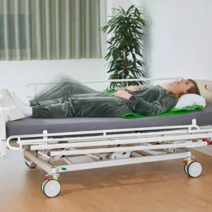 Person lying on an adjustable hospital bed with a gray outfit and green blanket in a clinical room with a plant nearby.
