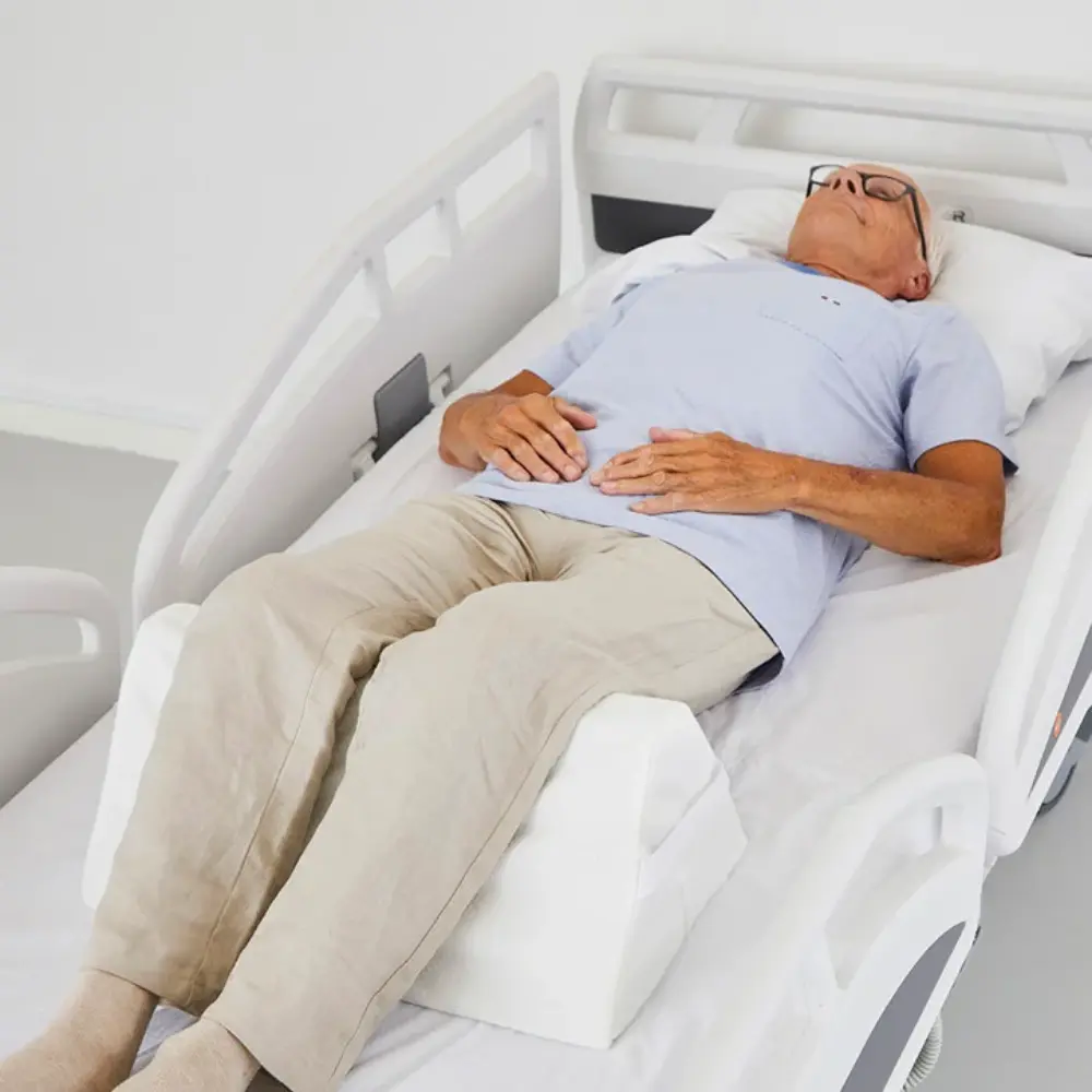 Older man resting in a hospital bed, hands on his abdomen, wearing a light blue shirt and beige pants with glasses on.