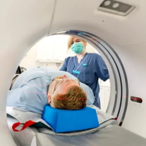 Patient lying on a CT scanner bed with head supported by a blue cushion as a nurse in scrubs and a mask stands nearby during the scan.