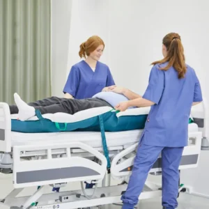 Two nurses in blue scrubs assist a patient lying on an adjustable hospital bed with side rails in a clinical room.