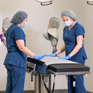 Two medical professionals in blue scrubs and masks position a sterile sheet on an operating table during a procedure.