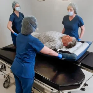 Patient lying on a hospital stretcher while three healthcare workers in blue scrubs and masks assist with the transfer.