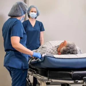 Two healthcare workers in blue scrubs and masks stand beside a patient lying on a wheeled hospital stretcher.