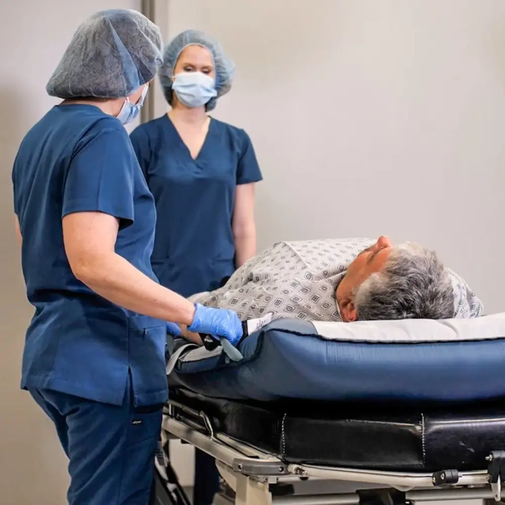 HoverMatt® T-Burg™ 5 - O Neill Healthcare Two healthcare workers in blue scrubs and masks stand beside a patient lying on a wheeled hospital stretcher.