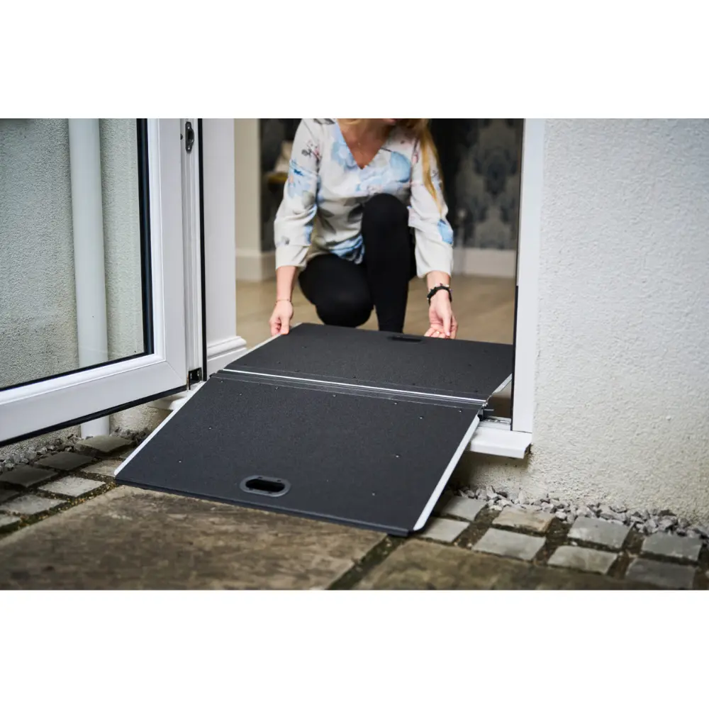 Doorline Butterfly Ramp 2 - O Neill Healthcare Person crouching to install a portable wheelchair ramp at a doorway outside a glass door.