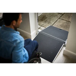 Wheelchair user seated in a blue jacket approaching a portable doorway ramp at an entryway.