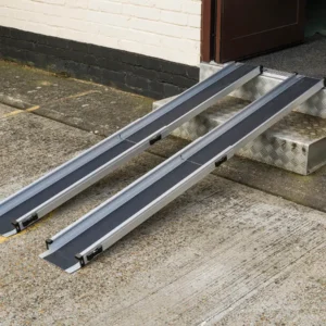 Two aluminum portable wheelchair ramps extending from a doorway onto a concrete surface for building access (dual ramps).